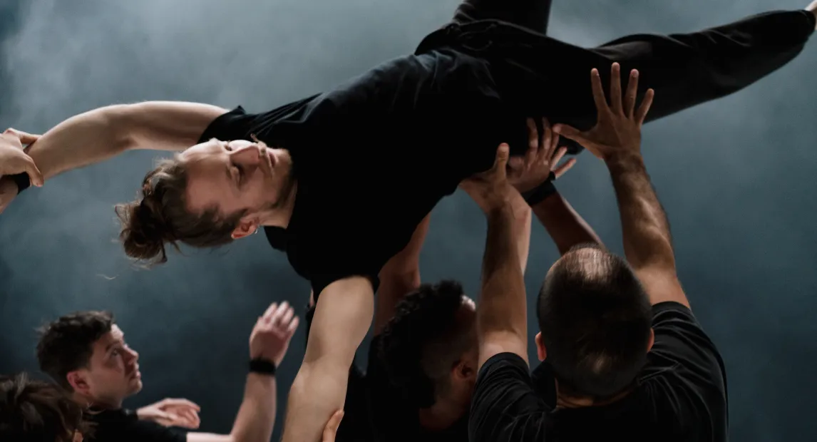 Un grupo de bailarines sostienen con sus manos en el aire a otro.